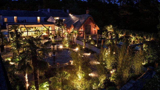 A well-lit courtyard with a thatched-roof building, surrounded by palm trees and a small pond. -  Le Petit Chateau & William de Percy (Newcastle-upon-Tyne)