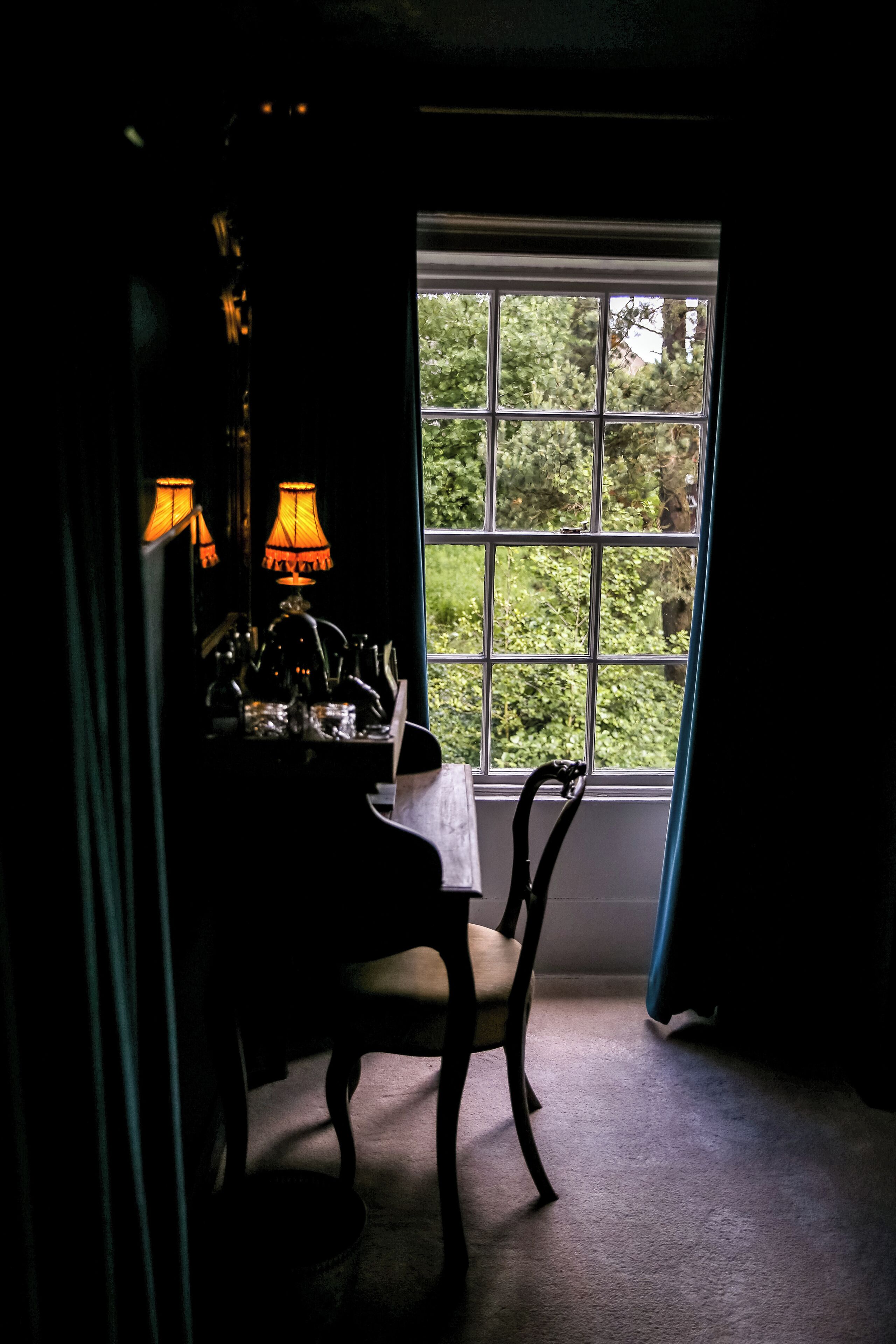 Ein schwach beleuchteter Raum mit Tisch und Stühlen, einem Fenster mit Blick auf Grünflächen und einer Lampe auf dem Tisch.