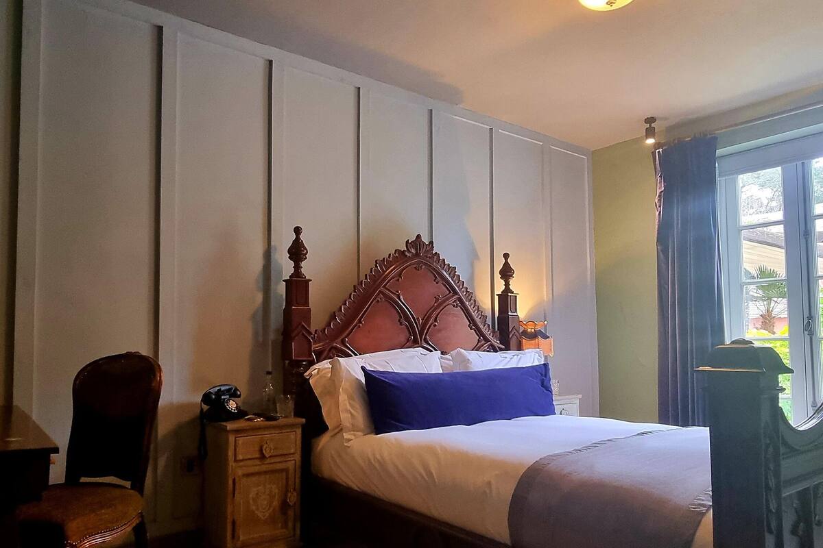 Une chambre à coucher avec un lit en bois, une table de chevet, une chaise, un bureau avec une bouilloire et une fenêtre avec des rideaux.