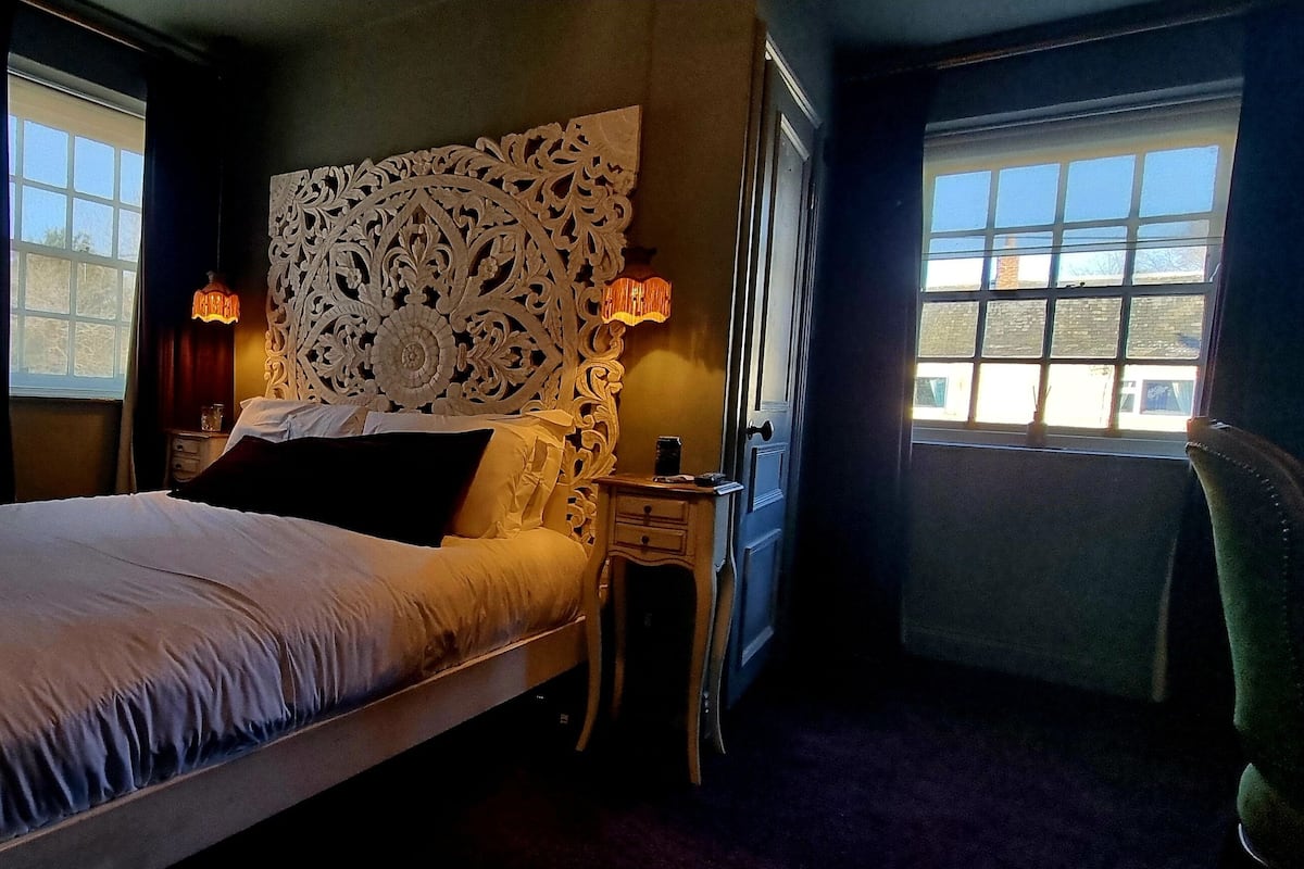 Une chambre à coucher comprenant un lit, une table de chevet, une tête de lit décorative, un miroir et deux fenêtres.