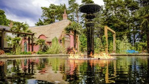 A pond with a fountain, surrounded by lush greenery and a building with a thatched roof. -  Le Petit Chateau & William de Percy (Newcastle-upon-Tyne)