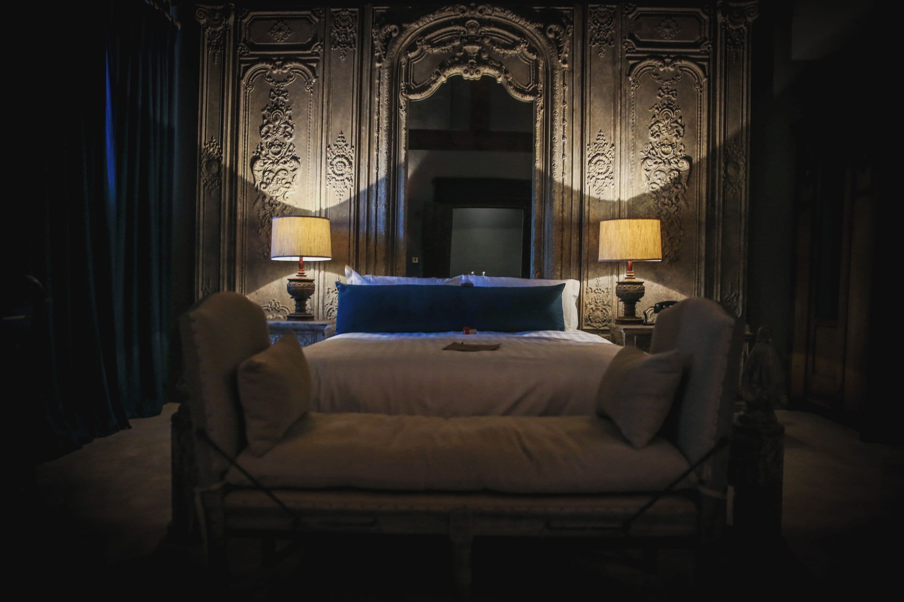 A dimly lit room with a large ornate mirror, a bed with blue and white linens, and two bedside lamps.