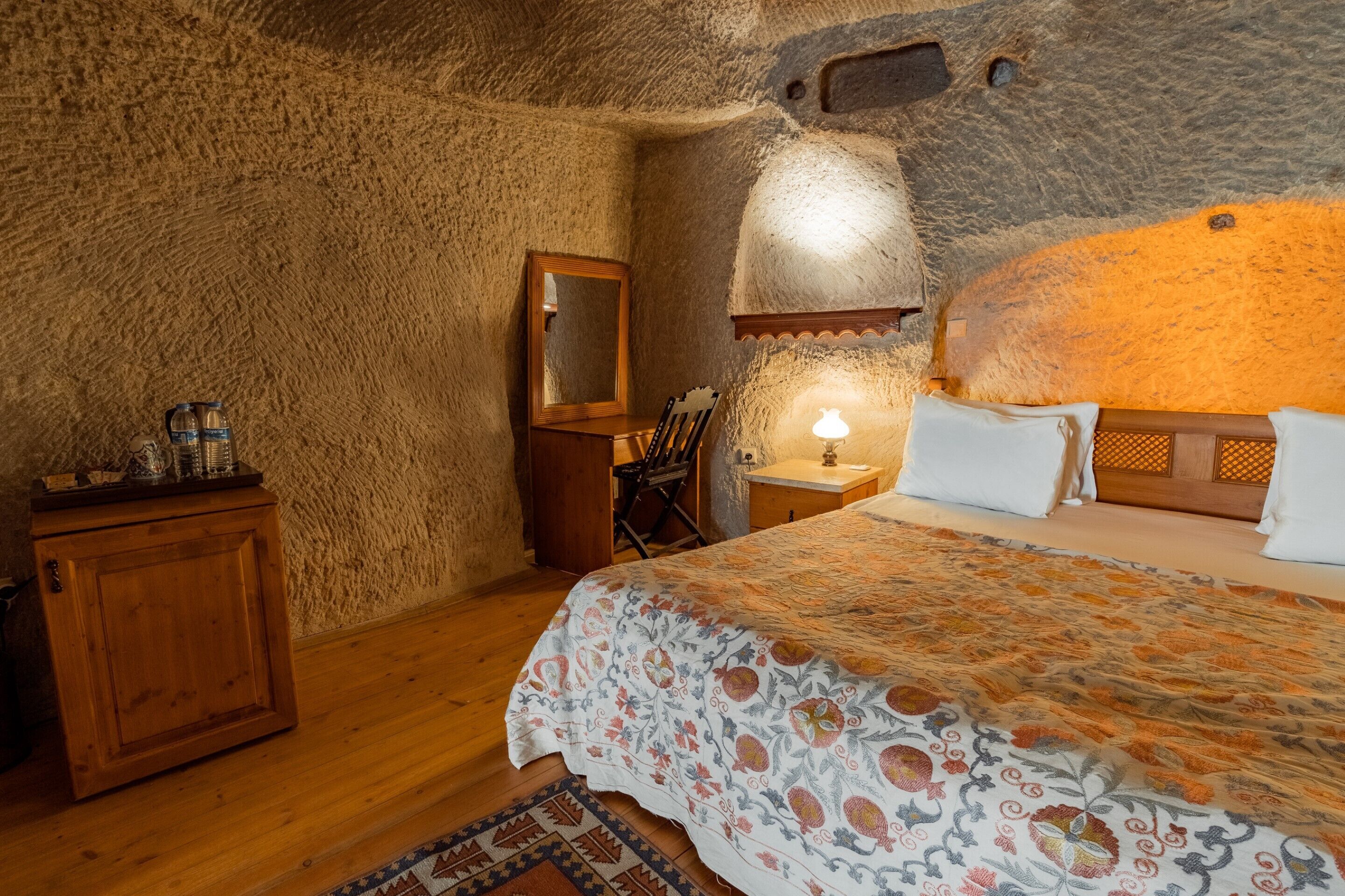 A bedroom with a stone wall, wooden furniture, a bed with patterned bedding, and a nightstand with a lamp.