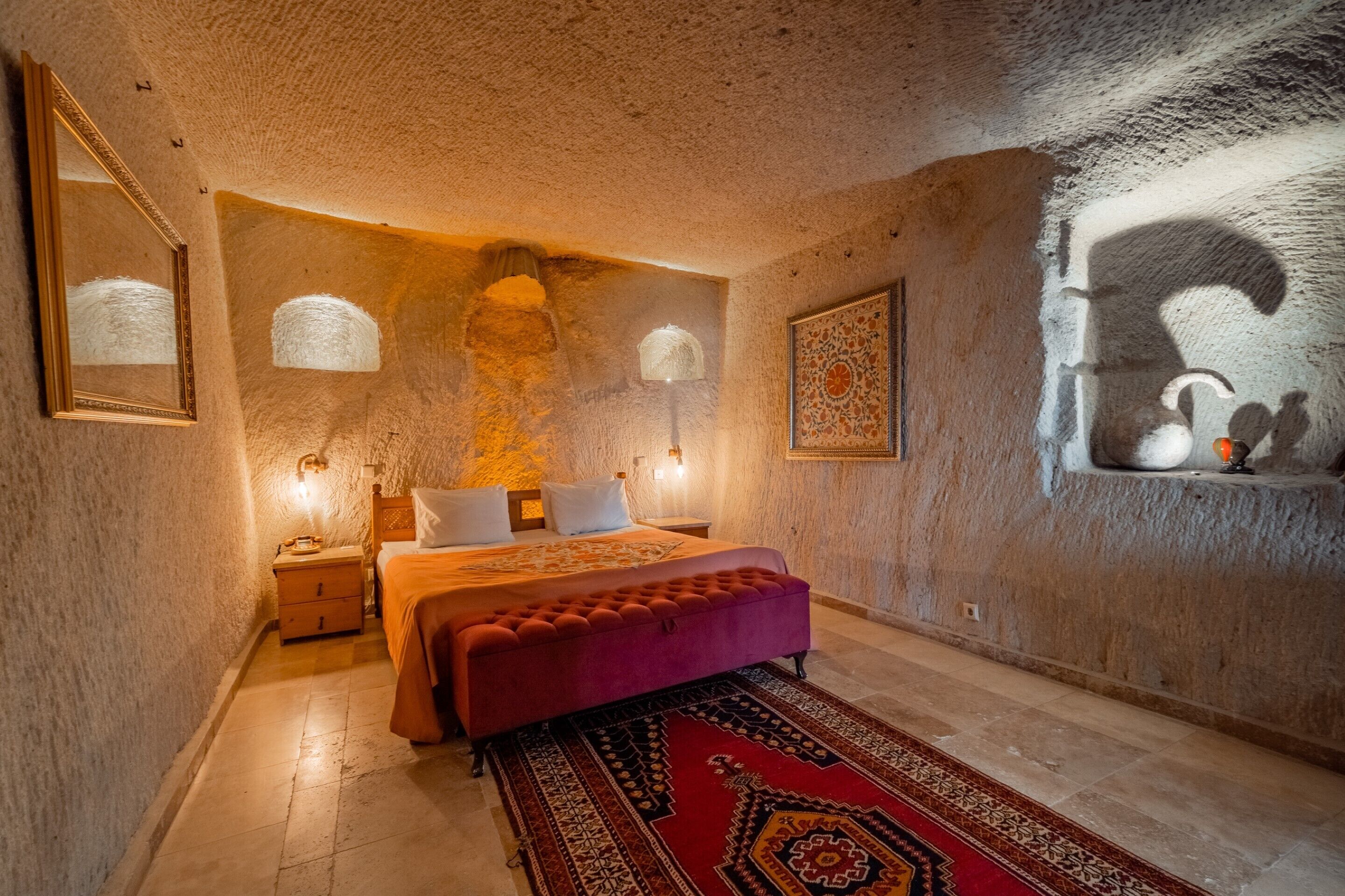 A bedroom with a large bed, a red ottoman, a patterned rug, and a wall-mounted light fixture.