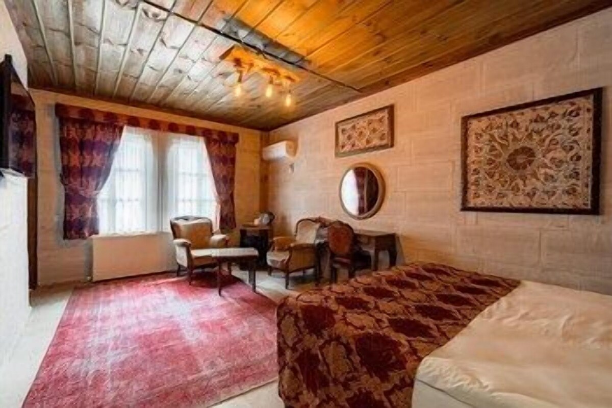 A room with a wooden ceiling, a bed, a desk with a chair, a round mirror, and two framed artworks on the wall.