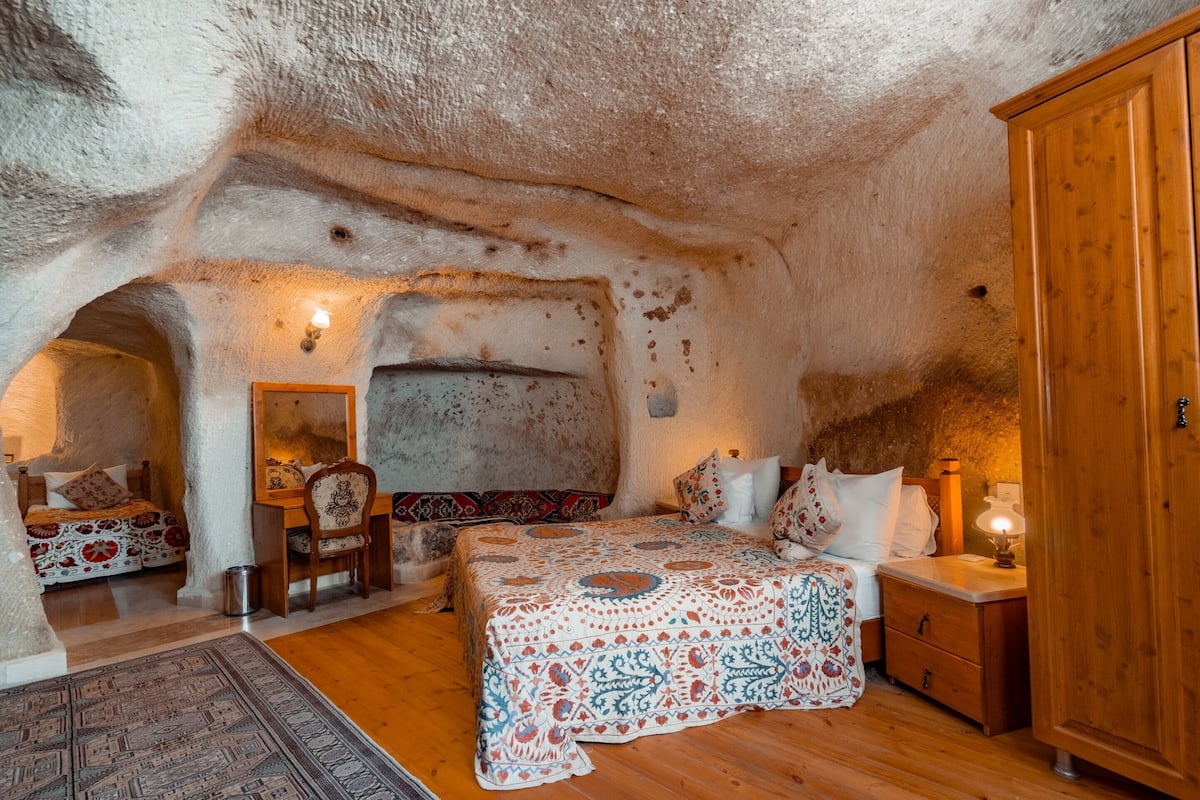 A cozy bedroom with a rustic cave-like ceiling, a wooden wardrobe, a bed with patterned bedding, and a small table with a lamp.