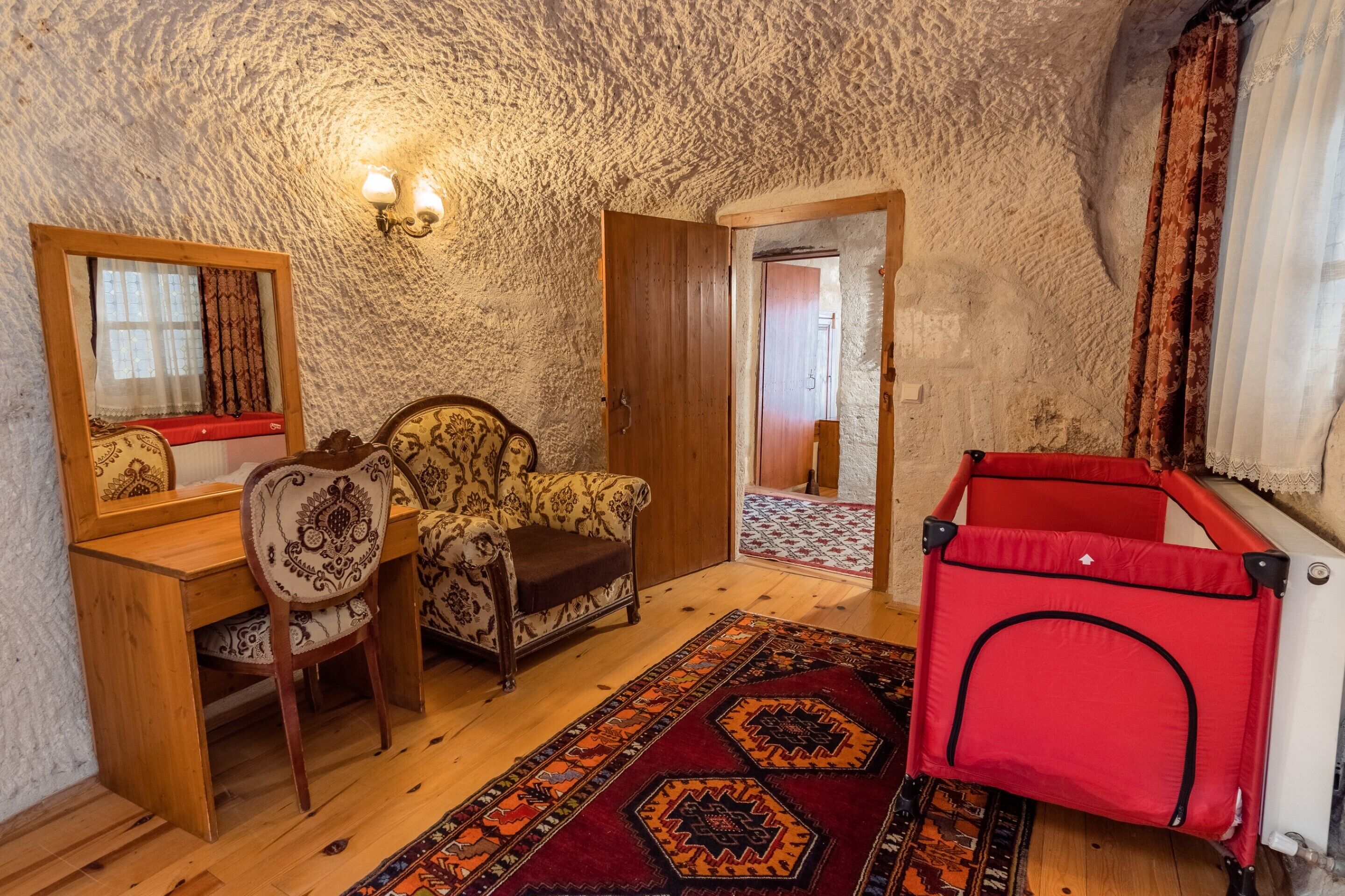 A cozy room with a wooden desk, a patterned armchair, a red playpen, and a mirror with curtains.