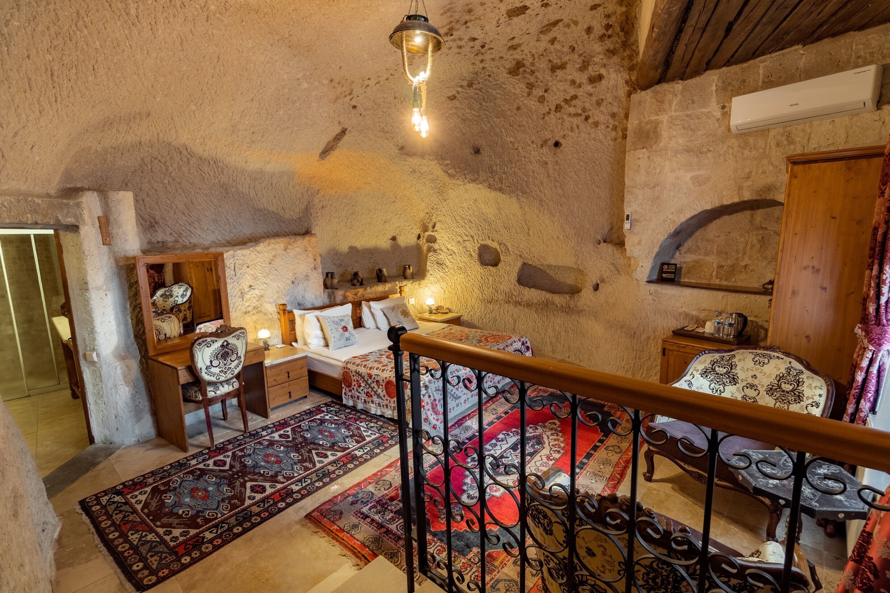 A rustic interior with a stone wall, a wooden desk, a bed with patterned bedding, and a red and black patterned rug.
