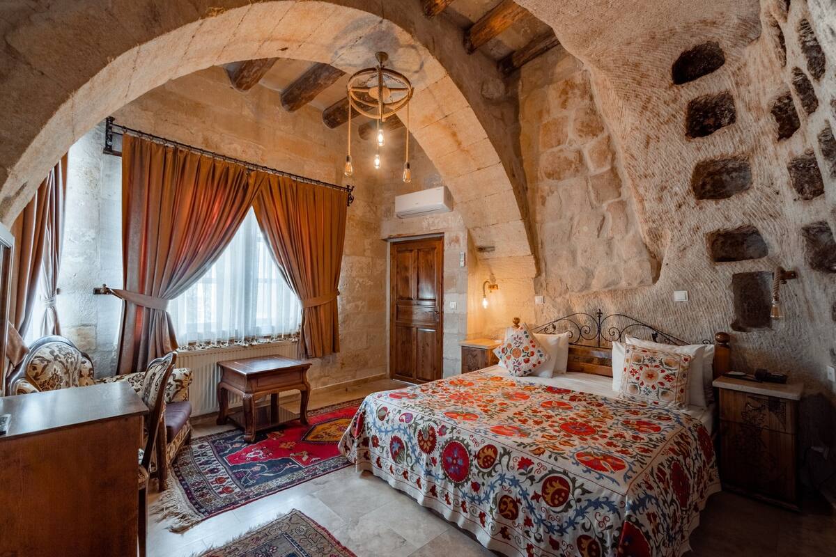 A bedroom with a large bed, a wooden desk, and a chair. There is a window with curtains, a chandelier, and a stone wall.