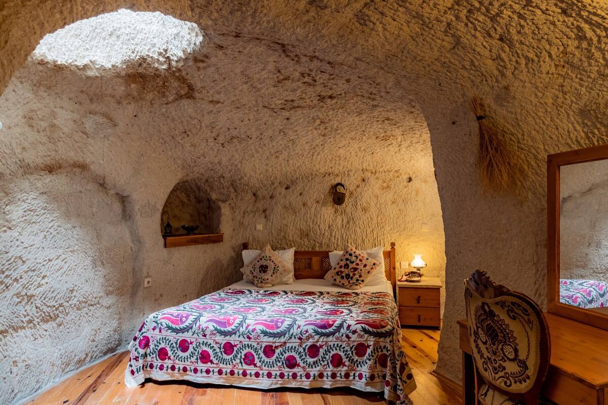 A rustic room with a stone wall, a wooden bed with patterned bedding, a nightstand with a lamp, and a mirror on the wall.
