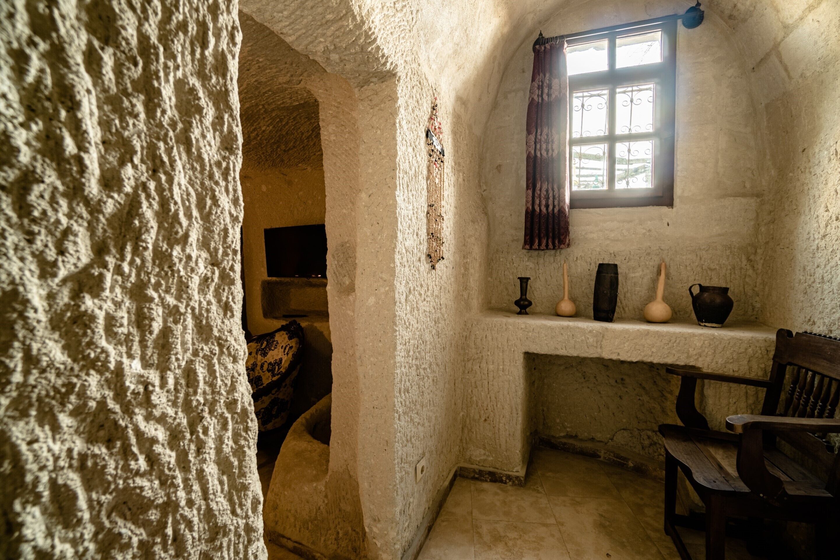 A rustic room with a stone wall, a wooden bench, a window with a curtain, and a shelf with vases.