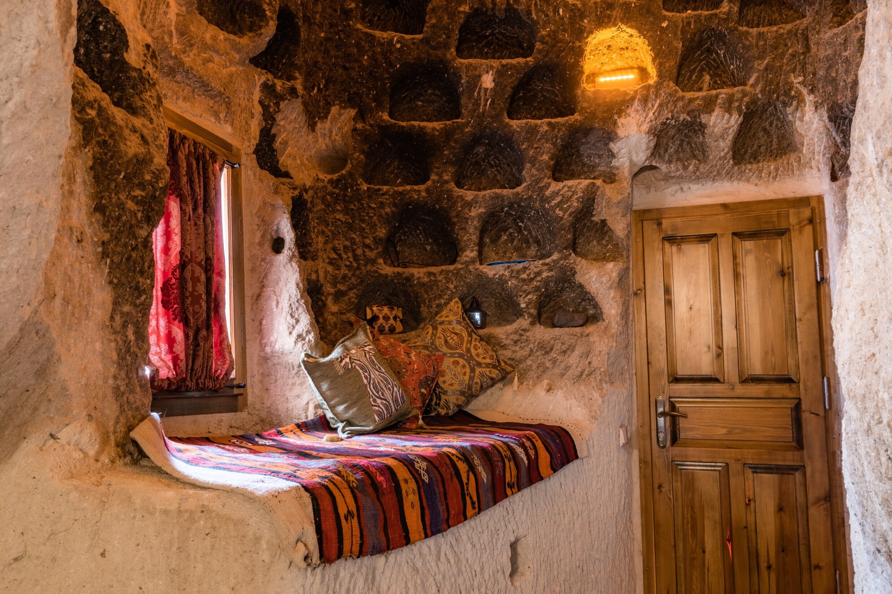 Una stanza con pareti in pietra, una porta di legno, un letto con una coperta colorata e una finestra con tende rosse.