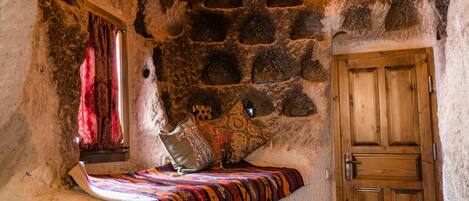 Una stanza con pareti in pietra, una porta di legno, un letto con una coperta colorata e una finestra con tende rosse.