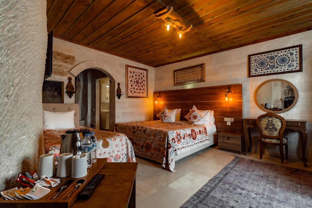 A cozy hotel room with two beds, a wooden desk, a mirror, and a stone wall.