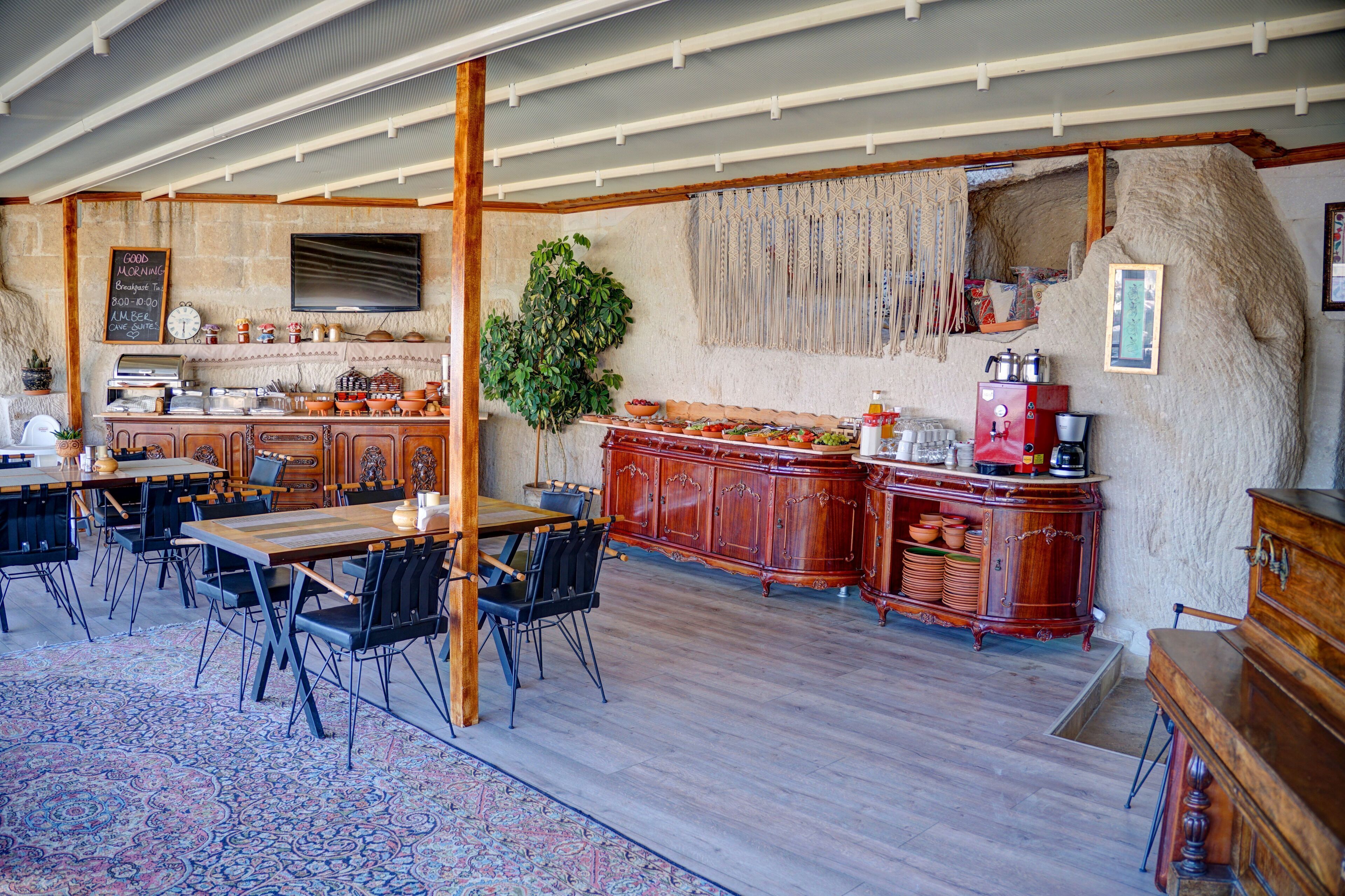 A rustic dining area with wooden tables and chairs, a large plant, and a traditional kitchen with wooden cabinets and a stone wall.