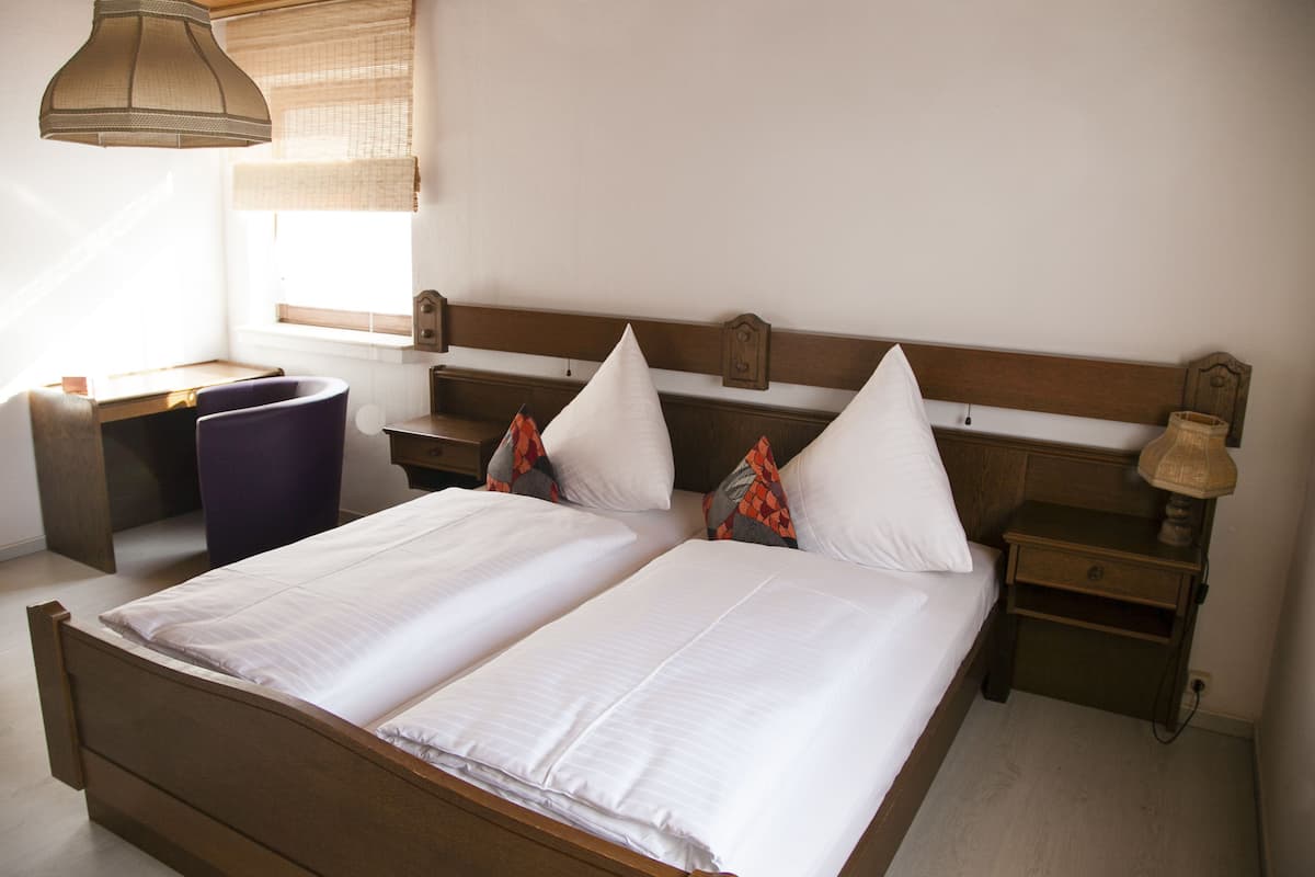 Una cama doble con ropa de cama blanca, cabecera de madera, dos mesitas de noche con lámparas, un escritorio y una silla.