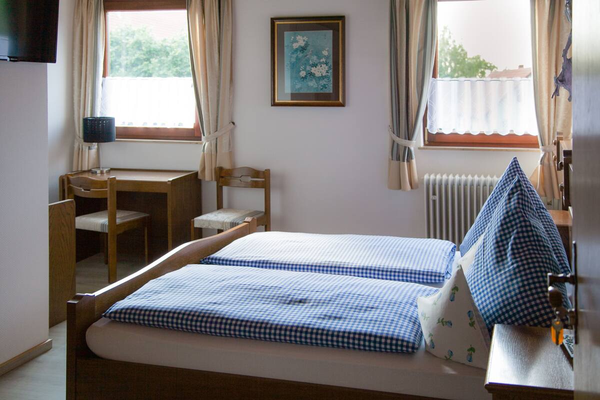 Un dormitorio con una cama, un escritorio y una silla. Se ve una ventana con cortinas, un radiador y un cuadro en la pared.