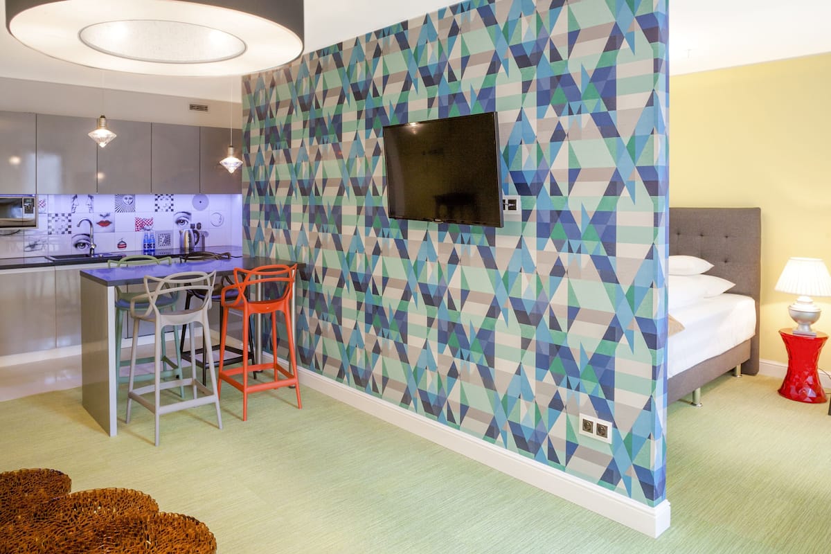 Ein modernes Hotelzimmer mit Wand im geometrischen Muster, einem flachbildfernseher über einem Essbereich, einem Bett mit grauem Kopfteil und einem Nachttisch in Rot.