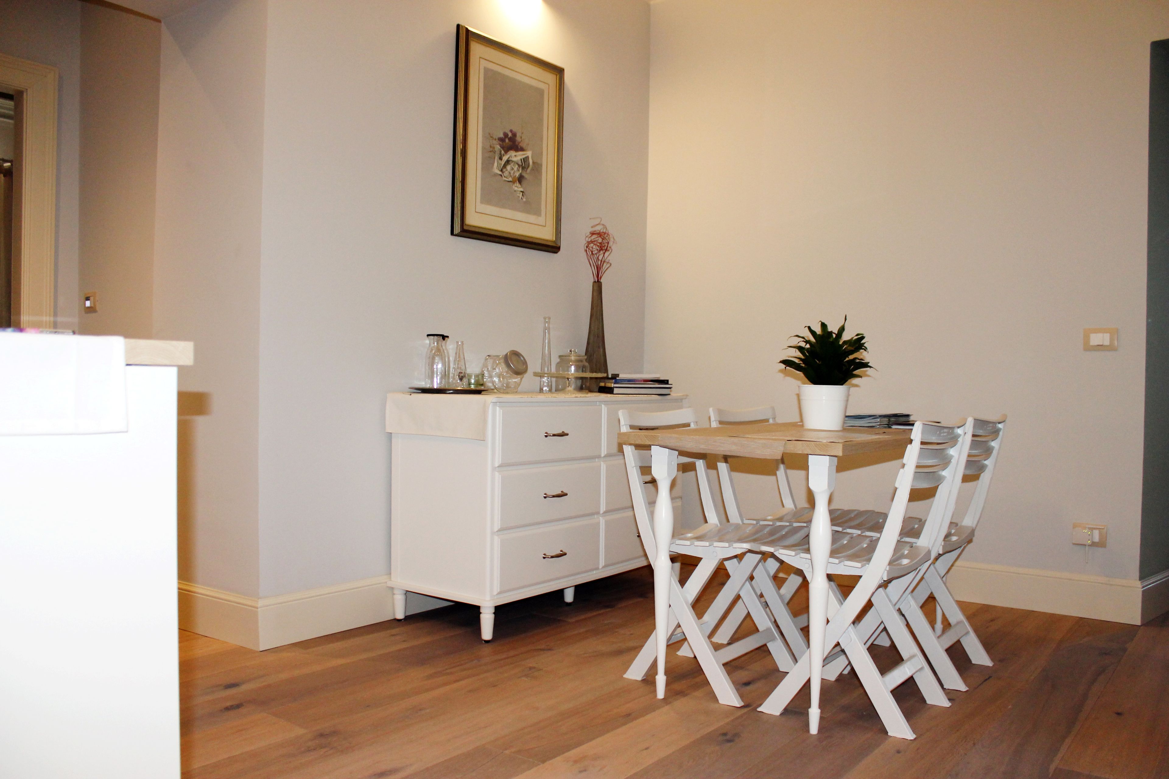 A dining area with a wooden table and white chairs, a white dresser with drawers, a vase with flowers, and a framed picture on the wall.