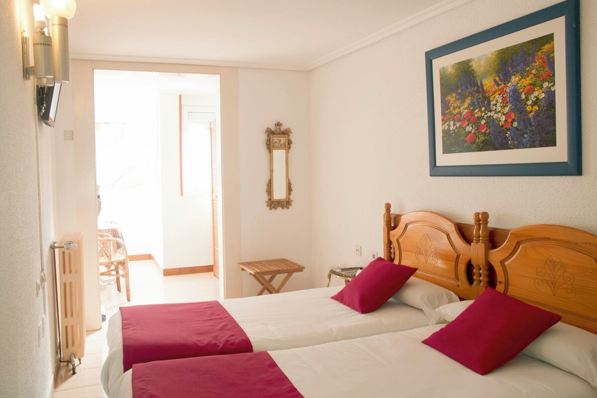 A hotel room with two beds, a wooden headboard, a framed floral painting, a small table, and a chair.