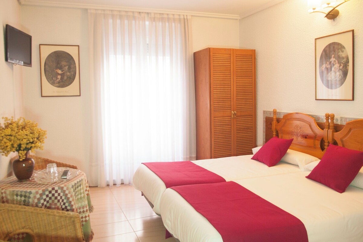 A hotel room with a bed, a wardrobe, a small table with a vase and a glass, and a wall-mounted TV.