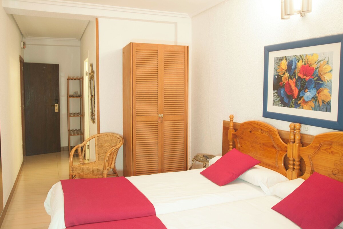 A hotel room with a bed, a wardrobe, a chair, a small table, and a painting on the wall.