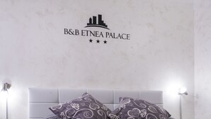 A hotel room with a bed, two patterned pillows, a nightstand with a lamp, and a wall with the hotel's logo and three stars. - B&B Etnea Palace (Catania)