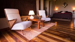 A room with two white armchairs, a small wooden table, a lamp, a bed with a dark cover, and a decorative wall piece. - El Soberbio Lodge (El Soberbio)