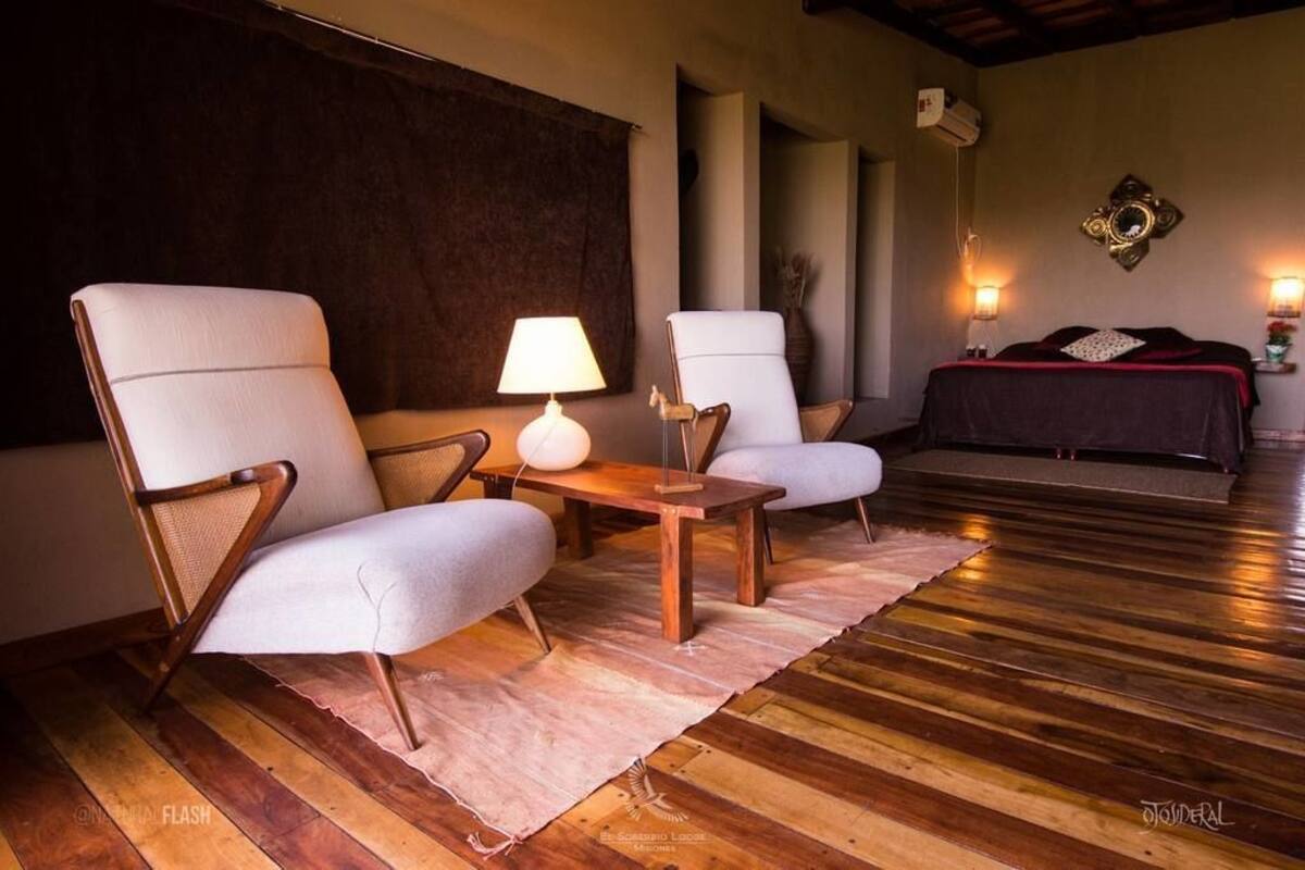 A room with two white armchairs, a small wooden table, a lamp, a bed with a dark cover, and a decorative wall piece.
