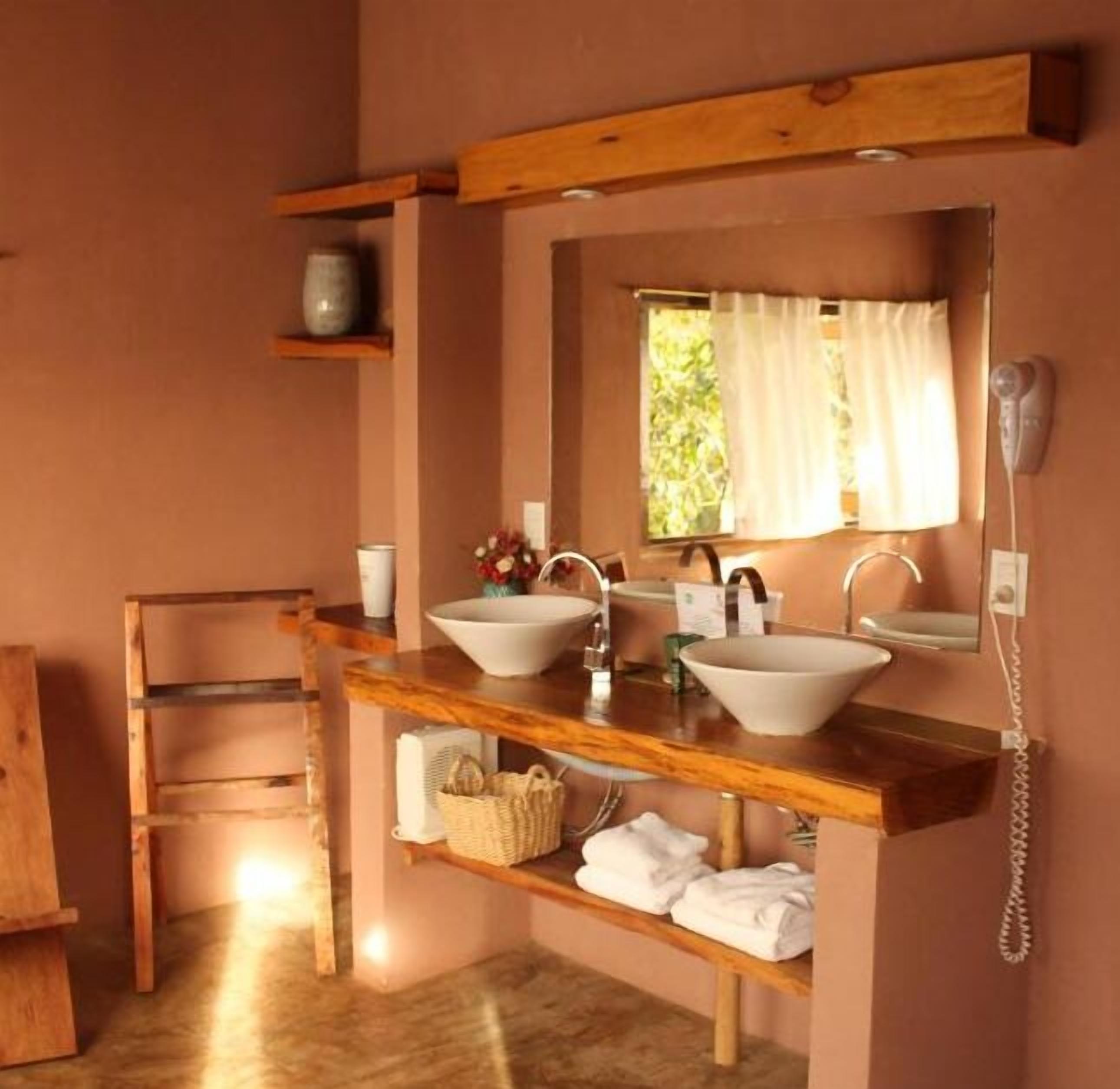 Un baño con estantes de madera, dos lavamanos, un espejo, un teléfono y una ventana con cortinas.