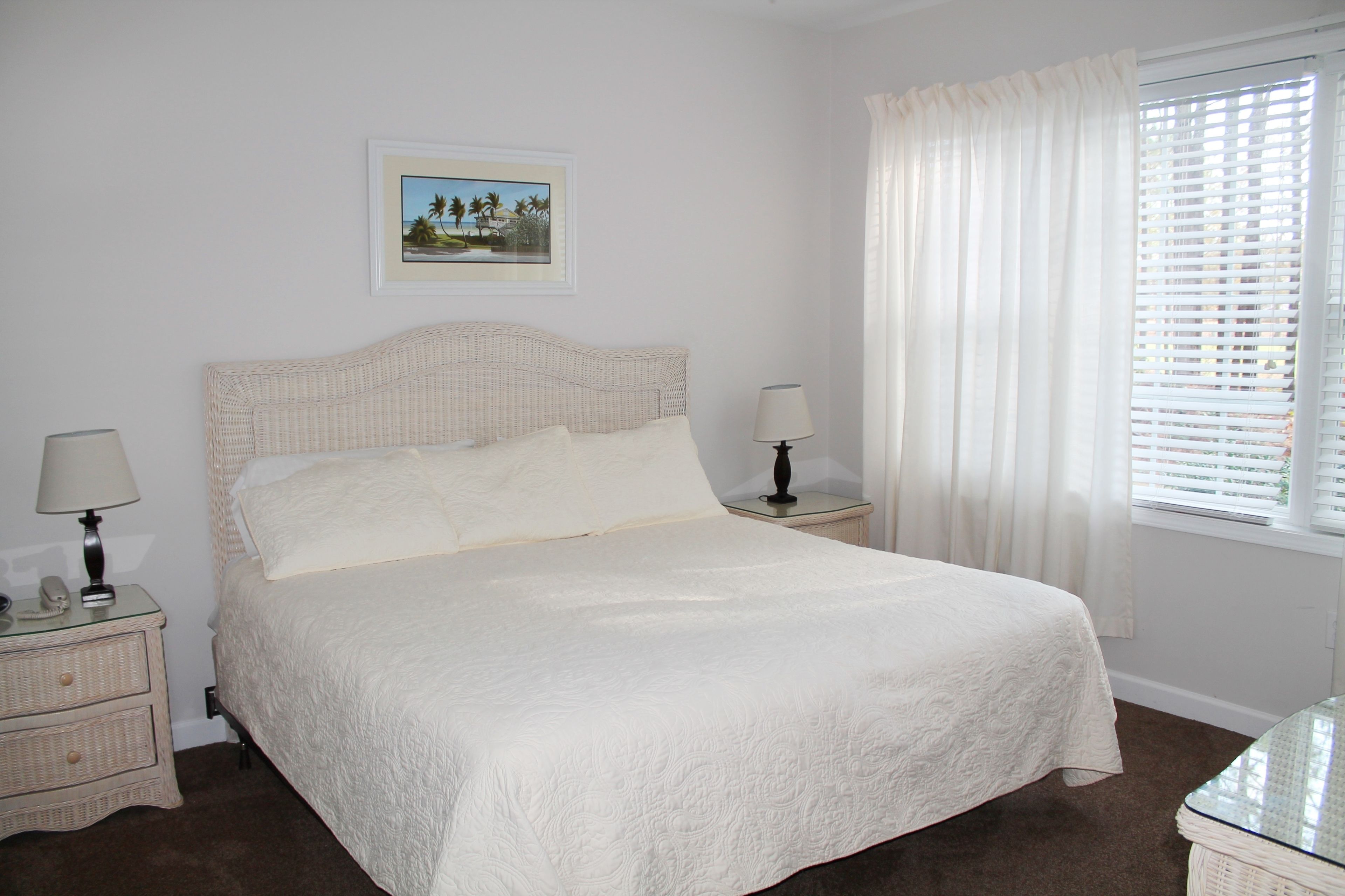 Una cama bien hecha con cubrecama blanco, dos mesitas de noche con lámparas, un cuadro enmarcado en la pared y una ventana con cortinas blancas.