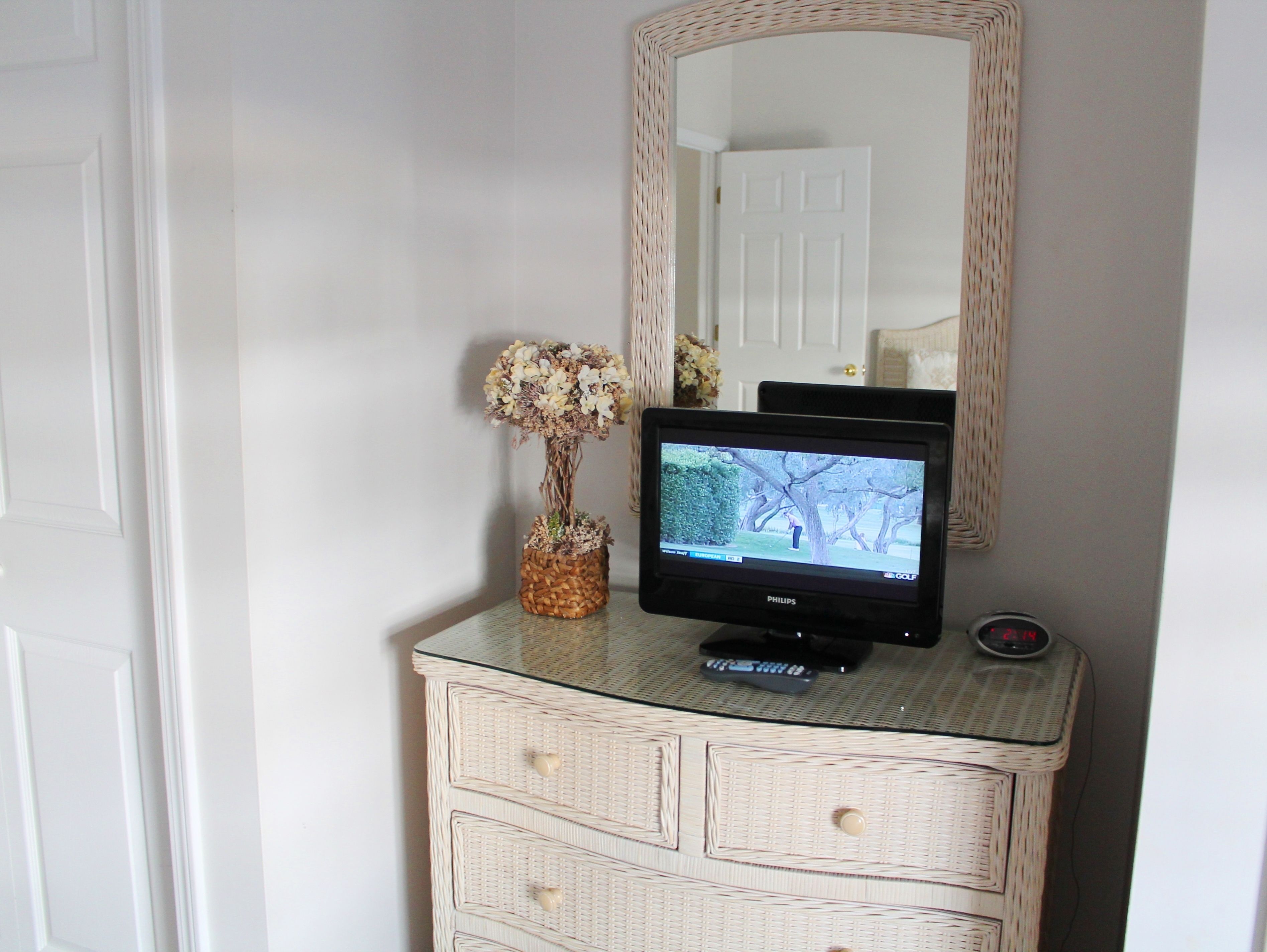 A television on a white dresser with wicker details, a vase with flowers, and a mirror above it.