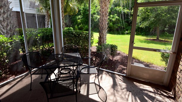 A covered patio with a table and chairs, a glass door leading to a garden with trees and grass. - 911 Willow Bend (North Myrtle Beach)