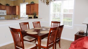 A dining room with a wooden table and chairs, a kitchen in the background, and a window with blinds. - 911 Willow Bend (North Myrtle Beach)