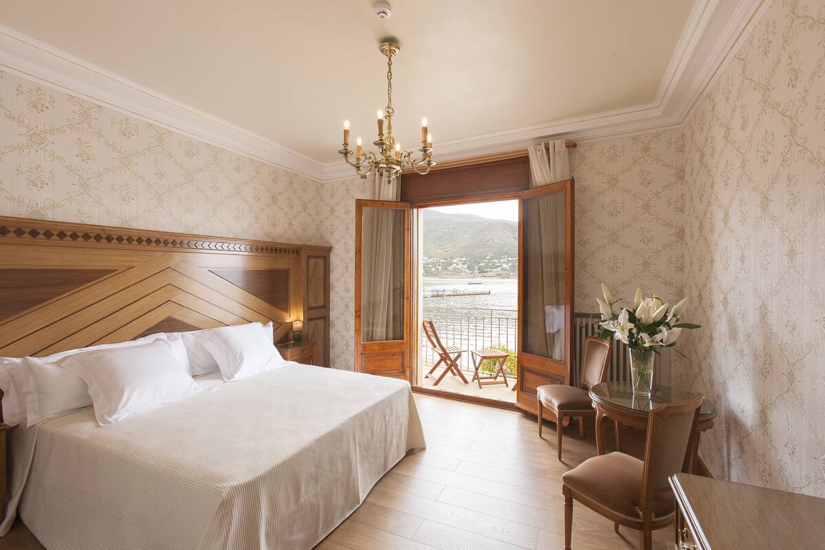 A hotel room with a large bed, a chandelier, a balcony with a view of a mountain, and a table with flowers.