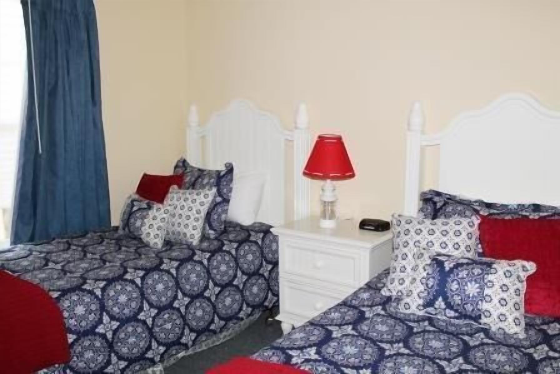 A double bed with a patterned duvet and pillows, a bedside table with a red lamp, and a window with blue curtains.