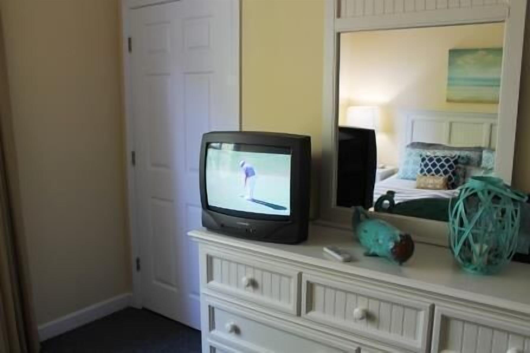 A television on a dresser with a remote control, a teal decorative item, and a mirror reflecting a bed with pillows.