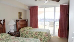 A hotel room with two beds, a ceiling fan, a nightstand with a lamp, and a view of the city through large windows.