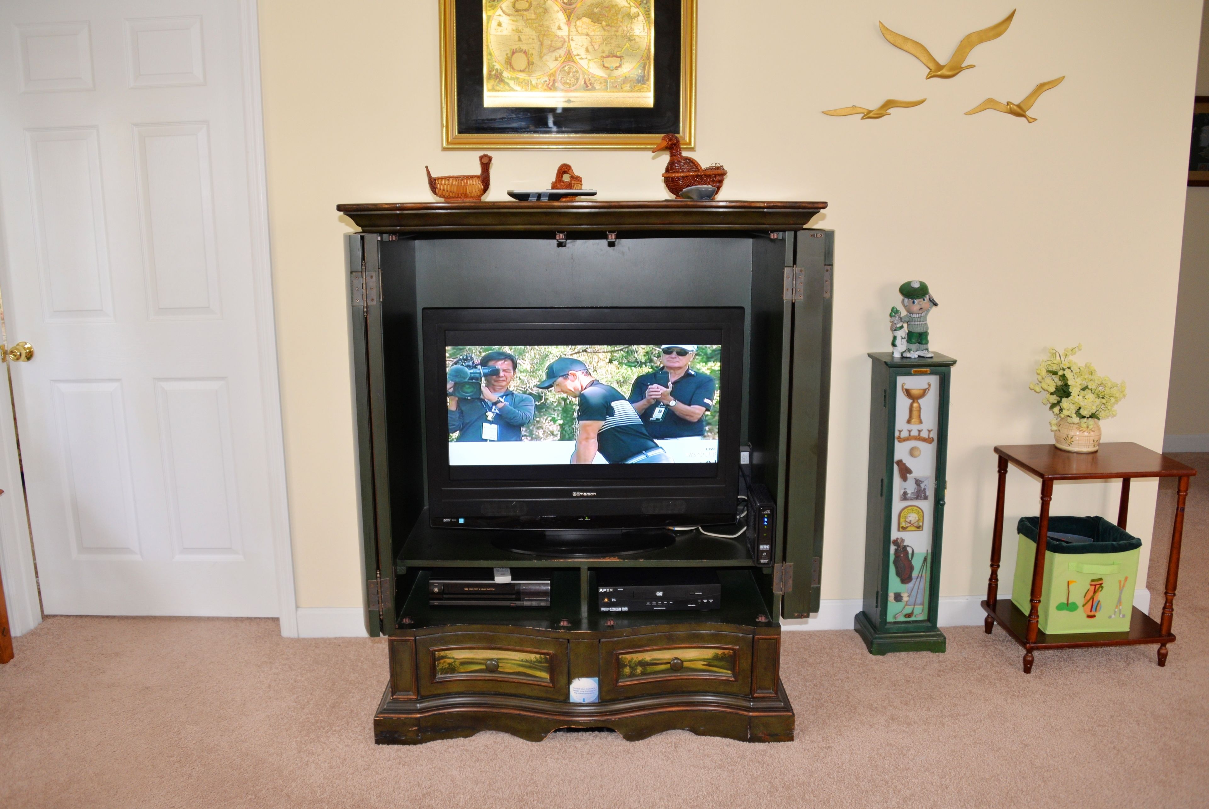 A living room with a television on a wooden stand, a small table with a green trash bin, and a decorative wall piece.
