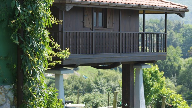 A wooden balcony house with a stone base, surrounded by lush greenery and a mountainous backdrop.