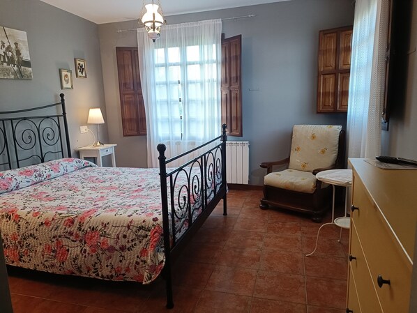 A bedroom with a bed, a chair, a dresser, a nightstand, a lamp, and a window with curtains.