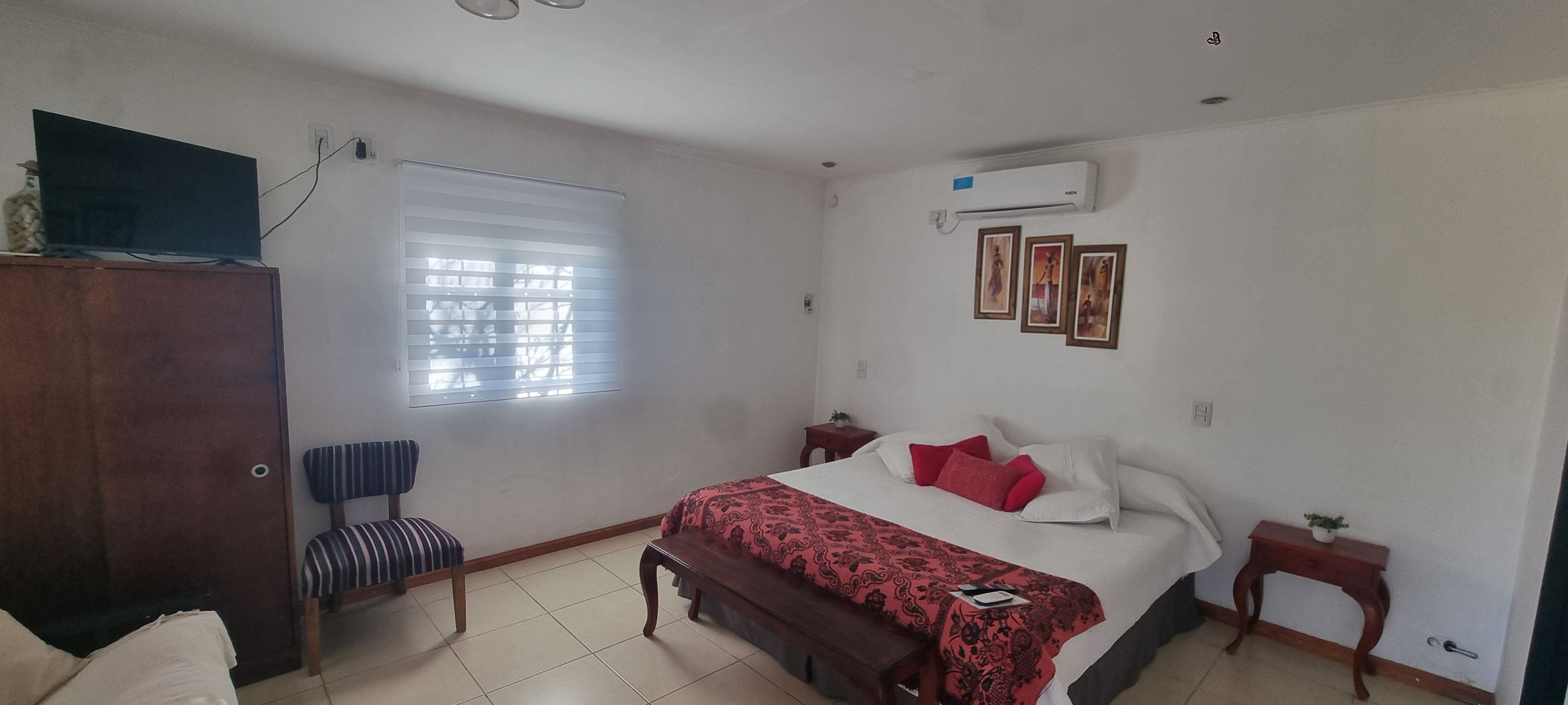 Un dormitorio con cama, ventana con persianas, mesita de noche, una pequeña mesa y un equipo de aire acondicionado.