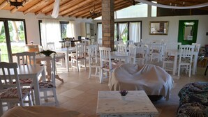 A spacious dining area with white tables and chairs, a central white table with a potted plant, and a bed covered with a white sheet in the foreground. - Finca Fisterra (Fray Luis Beltrán)
