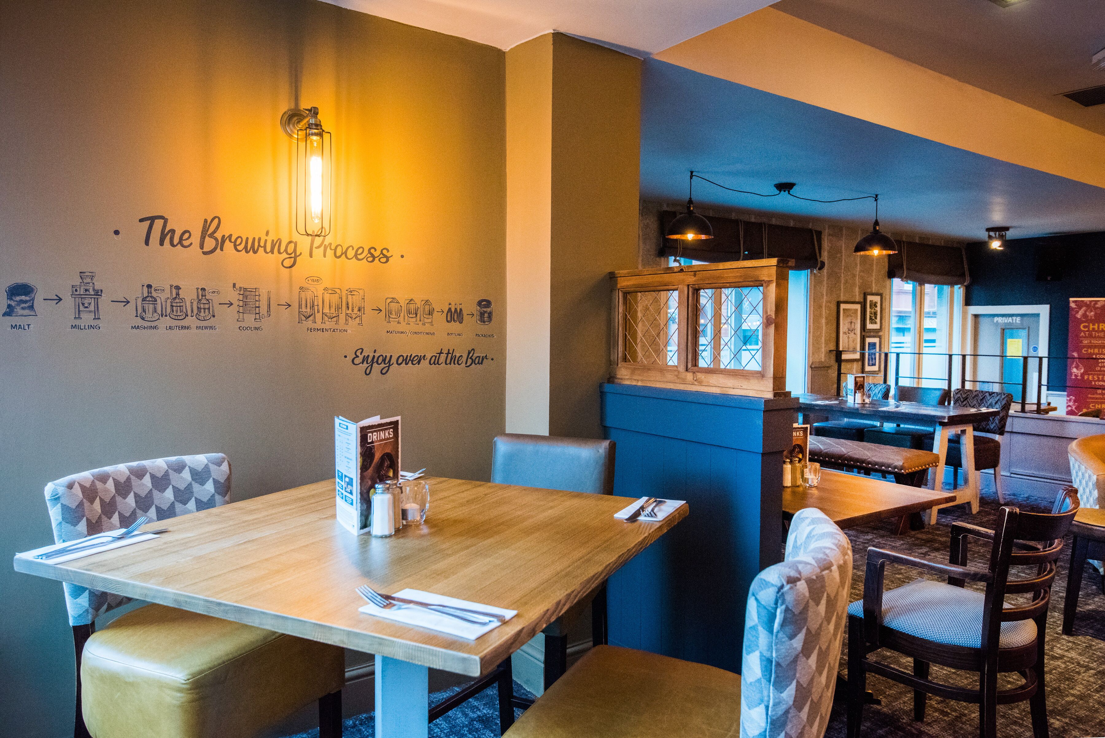 Interior de un restaurante con una pared que muestra el proceso de elaboración de cerveza, mesas de madera, sillas acolchadas e iluminación colgante.