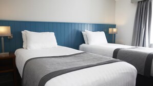 Habitación de hotel con dos camas, cada una con ropa de cama blanca y mantas grises. Hay lámparas de noche sobre mesitas de madera.