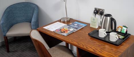 A hotel room with a wooden desk, a blue chair, a desk lamp, a coffee maker, two mugs, a tray with coffee pods, and a menu.