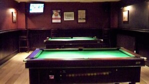 Una mesa de billar con superficie de fieltro verde, rodeada de paredes de madera oscura. Hay cuadros enmarcados en la pared y un televisor montado sobre la mesa.