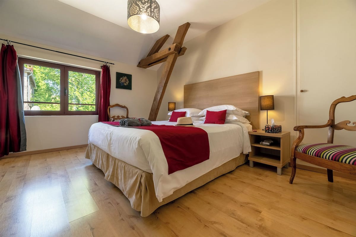 Un dormitorio con cama de madera, ropa de cama roja y blanca, una mesita de noche de madera, una silla y una ventana con cortinas.