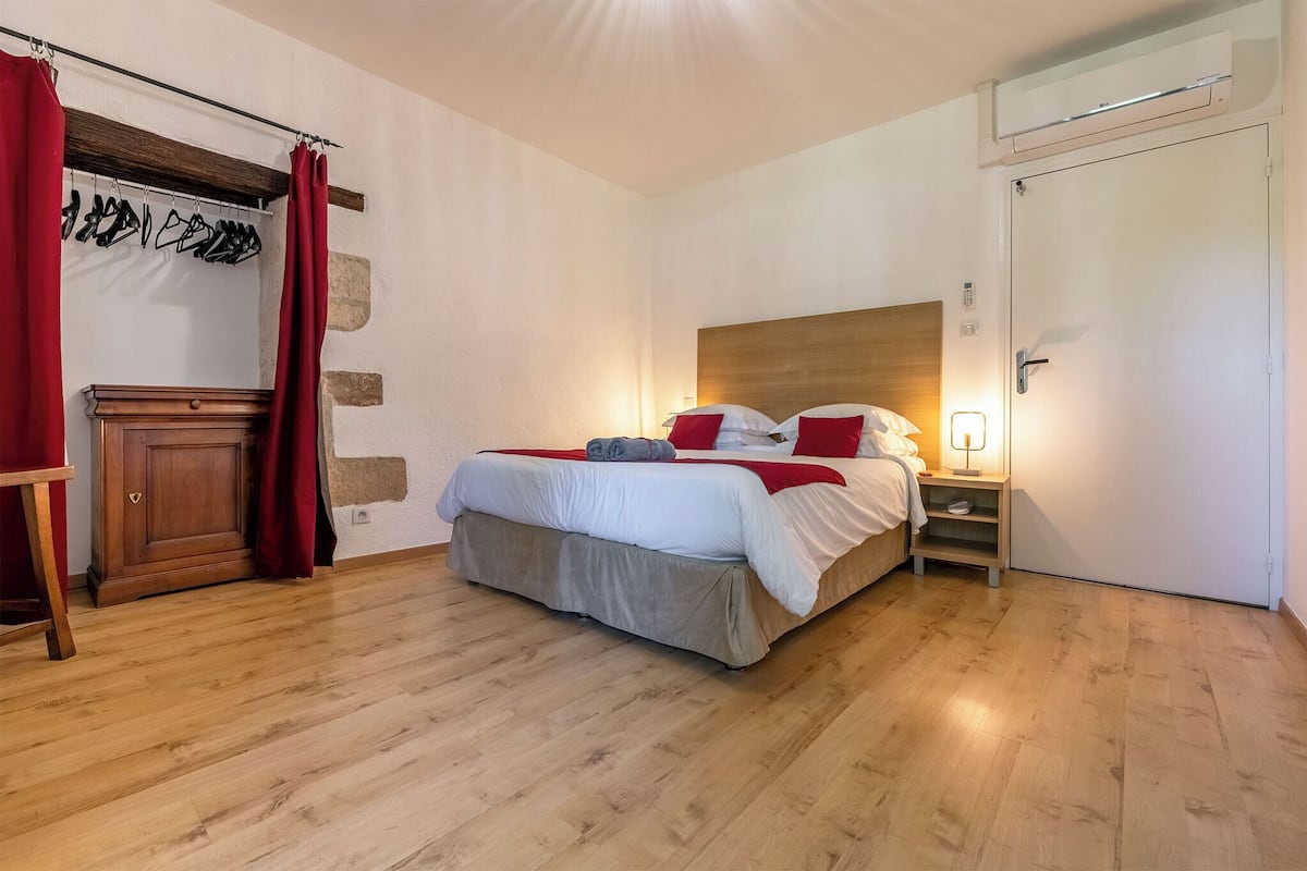 Un dormitorio con una cama de madera, cortinas rojas, una mesita de noche con lámpara y un armario.