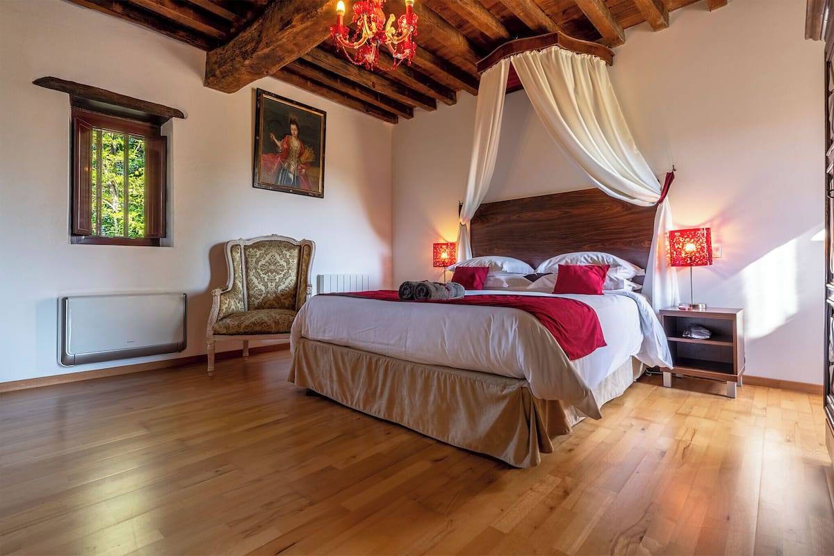 Un dormitorio con techo de madera, una cama con dosel, una silla, una mesita de noche y una ventana con vistas a la vegetación.